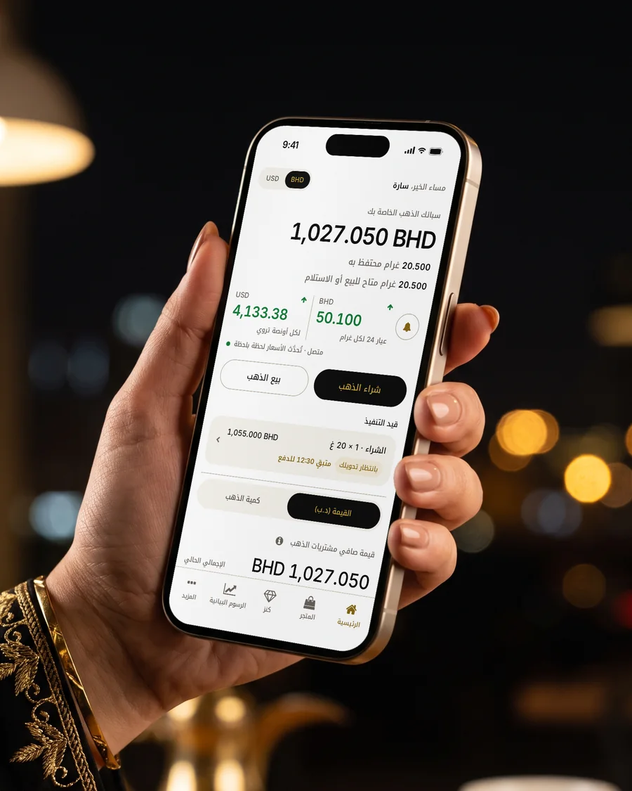 الشاشة الرئيسية لتطبيق MSS Gold على آيفون ذهبي تعرض سبائكك وطلباً قيد التنفيذ، فوق طاولة مقهى في المنامة ليلاً