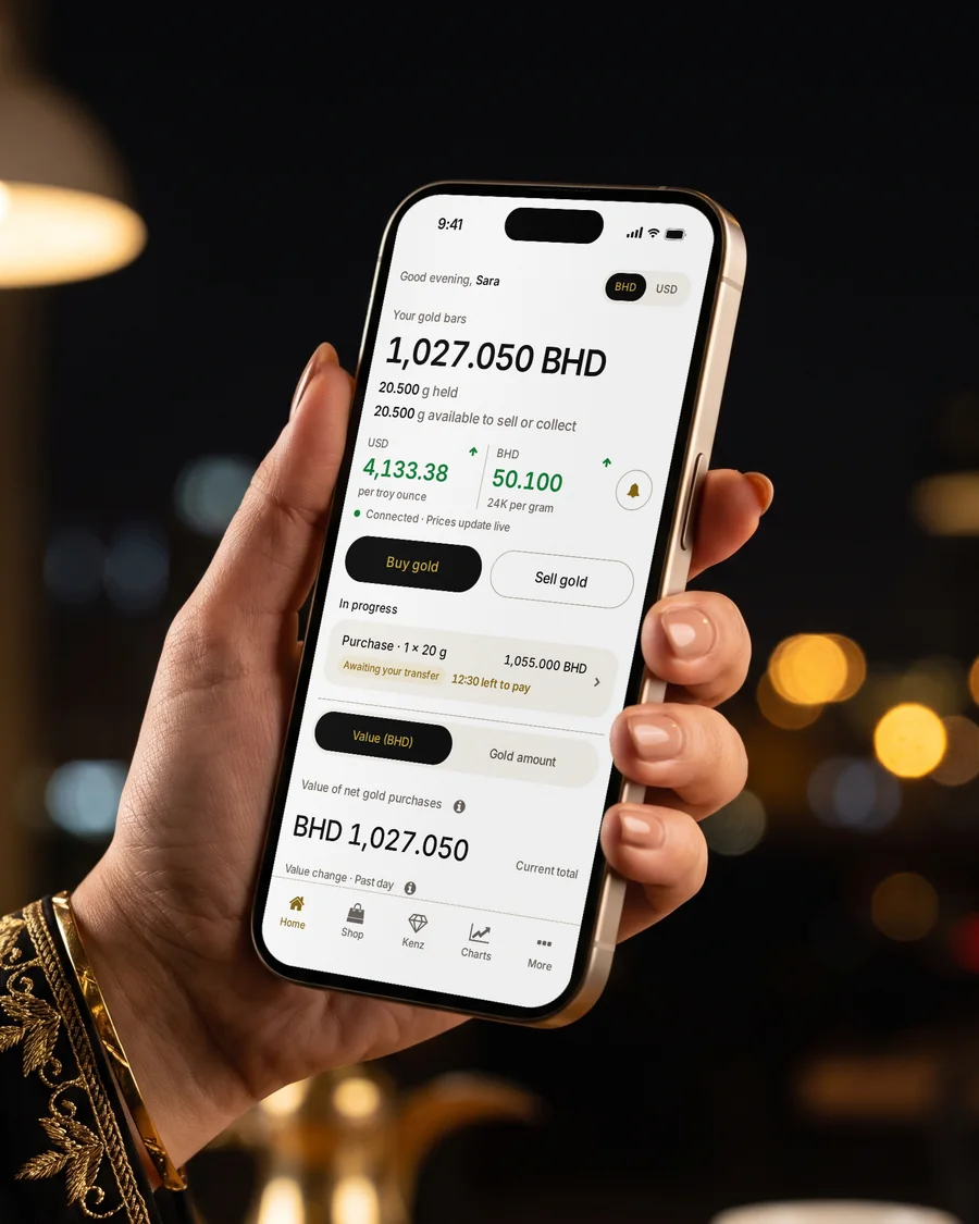 The MSS Gold App home screen on a gold iPhone, showing your gold bars and an order in progress, held over a café table in Manama at night